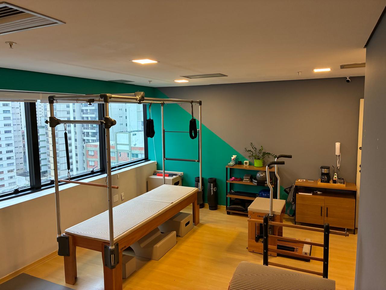 Ambiente do Voll Pilates Perdizes com aparelhos, parede verde e vista da cidade pelas janelas
